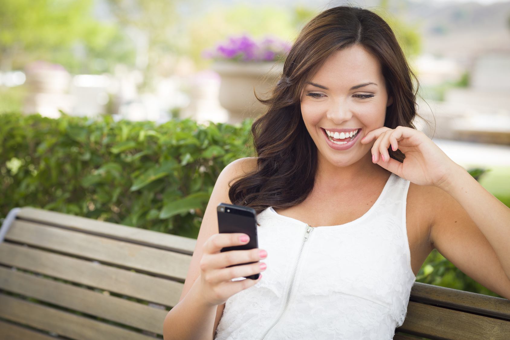Attractive Smiling Young Adult Female Texting on Cell Phone Outdoors on a Bench